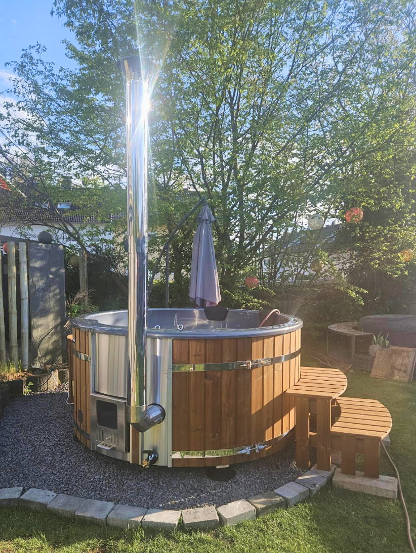 Wooden hot tub with stove and stairs in a garden with trees and a clear sky