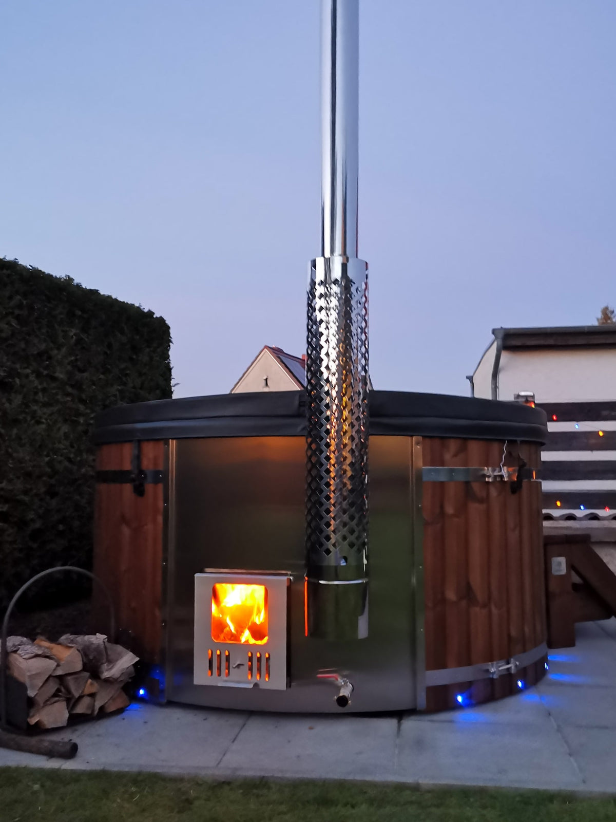 Outdoor view of wood fired hot tub with stainless steel stove, burning fire, wooden exterior panels, and tall chimney at dusk