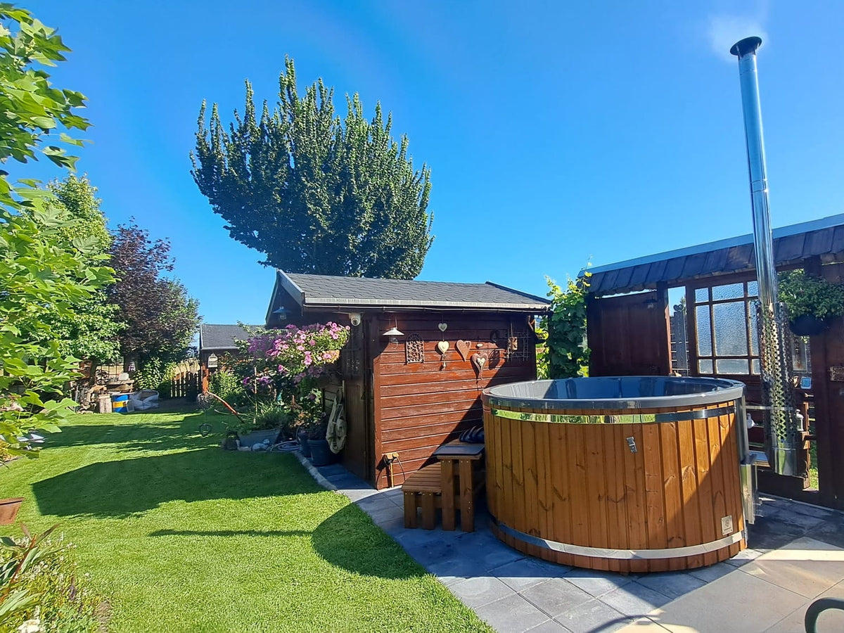 Outdoor view of round wooden hot tub with chimney next to wooden shed and green garden under clear blue sky