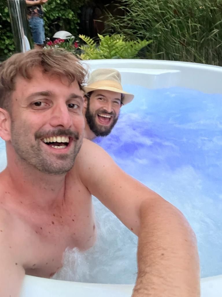 Two men smiling and relaxing in a bubbling outdoor hot tub surrounded by greenery in daylight
