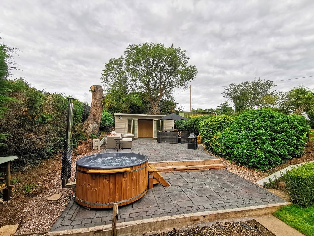 Outdoor view of a wooden hot tub with chimney on stone patio surrounded by greenery and garden seating area