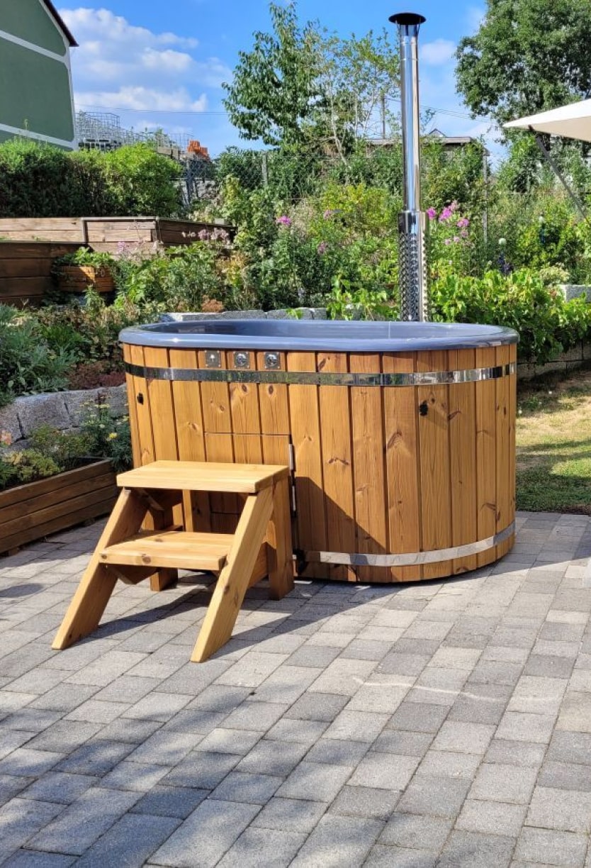 Outdoor view of wooden hot tub with metal bands, chimney pipe, and wooden steps on paved patio in garden setting