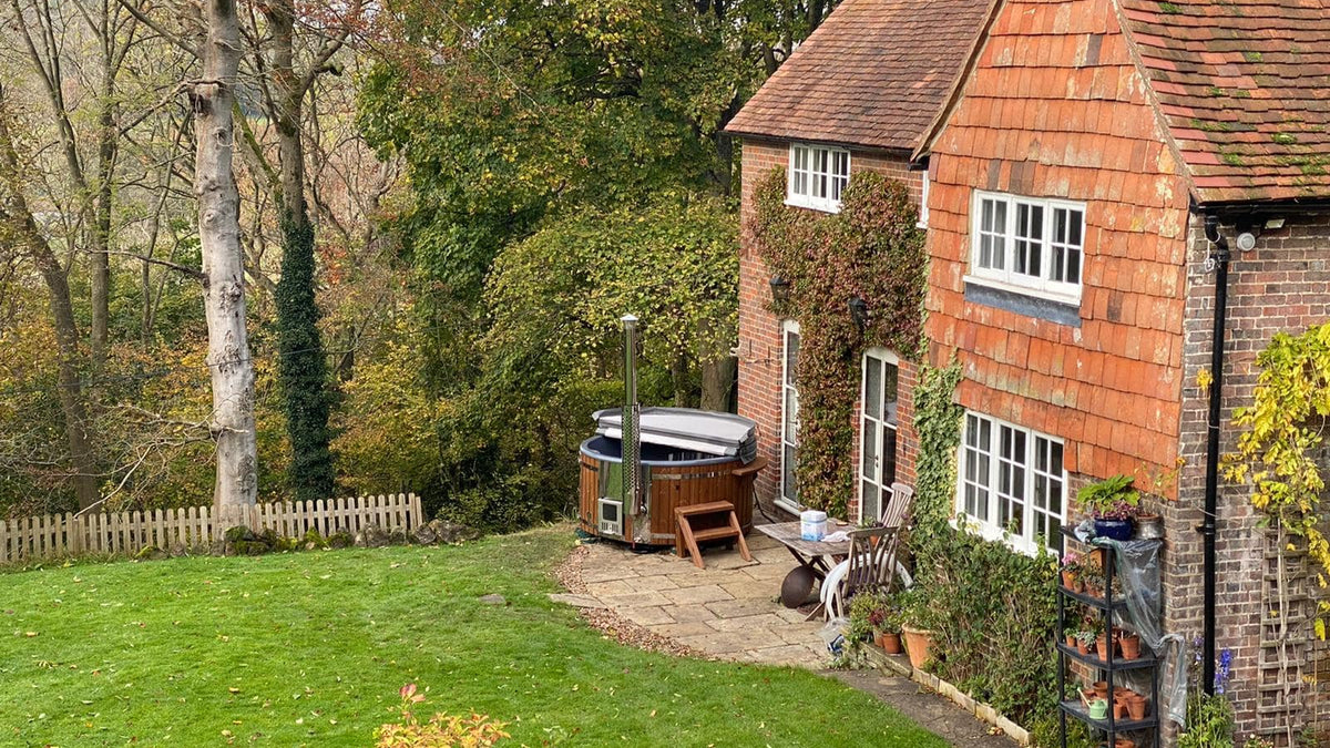 Outdoor view of a rustic brick house with a wood fired hot tub on a patio surrounded by green lawn and trees