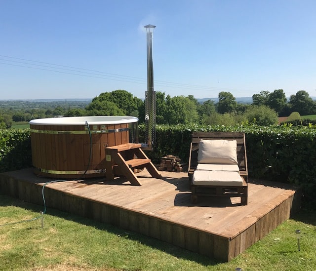 Outdoor view of round wood fired hot tub on wooden platform with small steps, chimney, and cushioned lounge chair