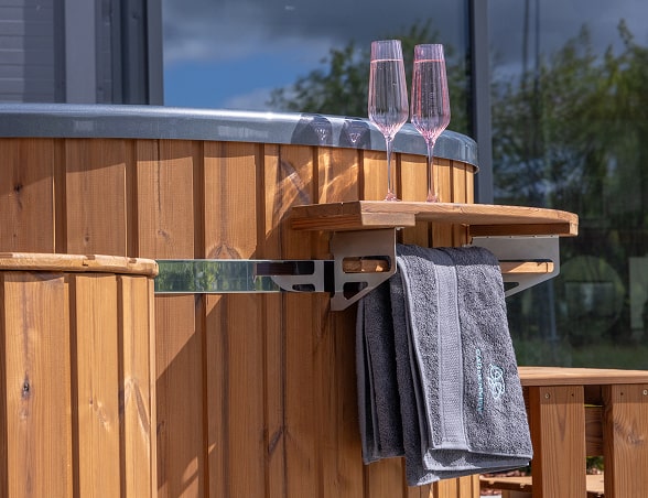 Close-up of wooden hot tub side with foldable tray holding two glasses and a hanging dark gray towel with embroidered logo