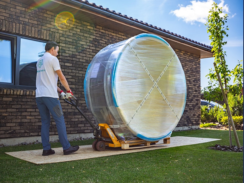 One man moving a wood-fired hot tub on a pallet truck and an OSB board to protect the lawn.