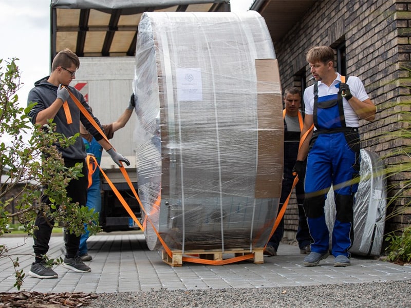 A packaged Gardenvity hot tub placed sideways on a wooden pallet and manhandled by four adults.